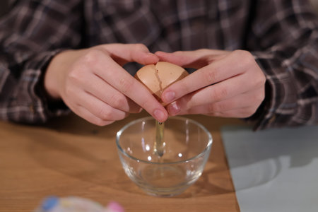 Close-up of hands cracking an egg over a glass bowl, preparing ingredients for baking on a wooden...