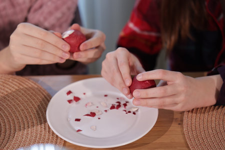 Two pairs of hands peeling red dyed Easter eggs over a white plate, with pieces of eggshell scattered on the plate.の写真素材