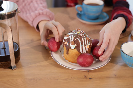 Hands arranging red dyed eggs around a decorated Easter cake topped with chocolate icing on a wooden table, with a French press and a blue cup nearby.の写真素材