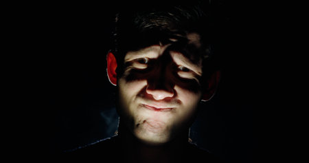 A man in darkness makes a bizarre and unsettling grimace, illuminated from below with dramatic lighting that creates deep shadows. A chilling and mysterious atmosphere.の写真素材