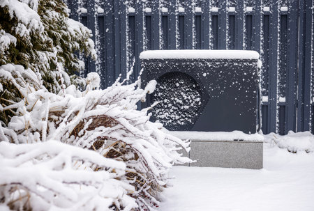Snow-covered outdoor heat pump unit beside frosted bushes and conifers against a gray metal fence in...