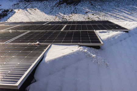 Solar panels partially covered with snow in a winter landscape, reflecting sunlight. The panels...