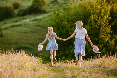 Middle-aged woman and young girl in blue dresses holding hands and walking through a scenic green field.の写真素材