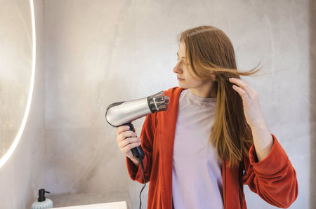 Young woman in a red robe drying her long hair with a hairdryer in a modern bathroom with soft lighting.の写真素材