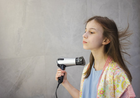 Teen girl in a floral robe uses a hairdryer in the bathroom, with her long hair dramatically blown by the warm airflowの写真素材