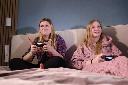 Two teenage girls sitting on a bed playing a video game, one focused with a serious look, the other...