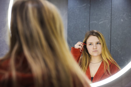 Young woman in a red robe stands in front of a glowing round mirror, thoughtfully examining her...