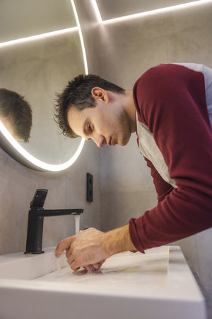 Young man practices hand hygiene at the sink, focusing on cleanliness and daily wellness in a bright and modern bathroom environment.の写真素材