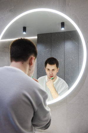 Young man in a light sweater brushes his teeth while looking in a round LED-lit mirror, maintaining his morning routine in a modern bathroom.の写真素材