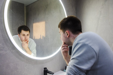 Man in a light sweater leans over a bathroom sink, thoughtfully examining his reflection in a round LED-lit mirror during his morning routine.の写真素材