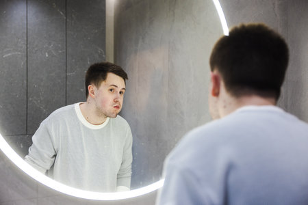 Man in a light sweater rinses his mouth while looking into a round illuminated bathroom mirror, finishing his morning dental routine.の写真素材