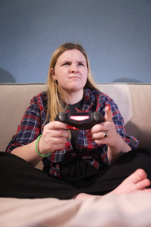 Focused teen girl plays video game at home, sitting cross-legged on a bed with a determined...