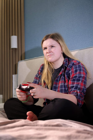 Teen girl sits barefoot on a bed with a wireless controller, showing a serious and competitive...