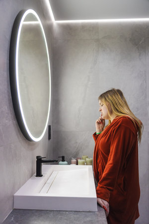 Young woman in a red bathrobe stands at the sink, brushing her teeth while looking at her reflection...