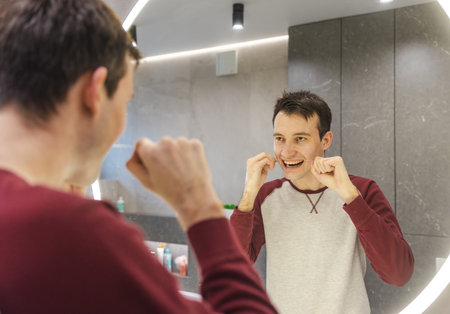 Young man smiles while flossing his teeth in front of a bathroom mirror, maintaining oral health and reinforcing a consistent hygiene routine.の写真素材
