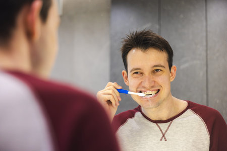 Smiling young man practices oral hygiene by brushing his teeth in front of the mirror, promoting...