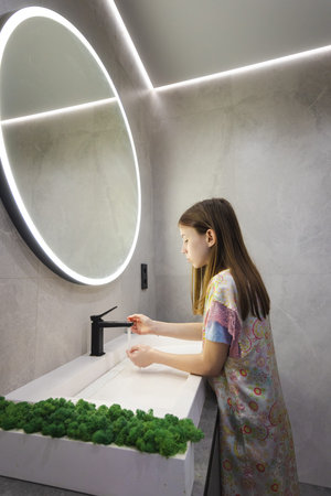 Young girl in pajamas finishes washing hands and turns off the faucet in a bright, stylish bathroom...
