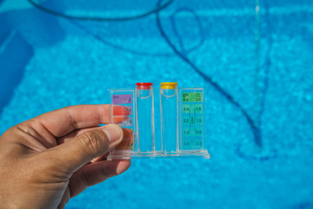 A person holds a clear pool water tester with pH and chlorine indicators above a sparkling blue swimming pool during regular maintenance on a sunny day.の写真素材