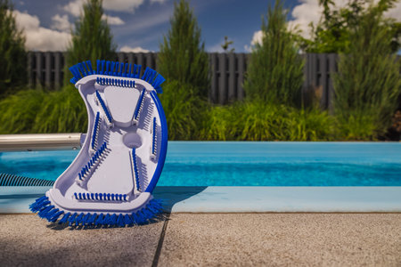 Curved pool vacuum head with blue bristles and hose connector resting on the poolside, ready for...