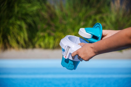 Hands placing chlorine tablet into pool dispenser with thermometer for clean and safe water...