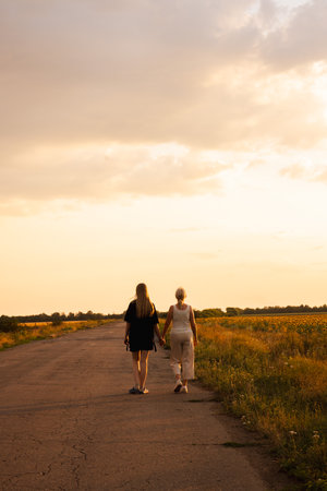 Two women walking hand in hand along a rural road at sunset surrounded by fieldsの写真素材