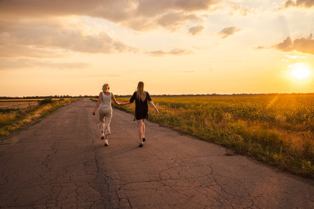 Two women walking hand in hand along a country road at sunset near blooming sunflower fieldsの写真素材