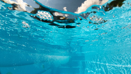 Underwater view of a backyard pool with steps, bubbles, and shimmering reflections, showing the surface tension and sky clouds from below the water.の写真素材