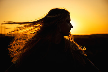 Teen girl in silhouette with flowing hair at sunset, gazing into the distance as warm golden light outlines her profileの写真素材