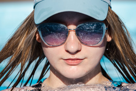 Teenage girl relaxing in a bubbling pool, wearing mirrored sunglasses and a pale visor cap, enjoying a sunny summer day with wet hair and a calm smileの写真素材