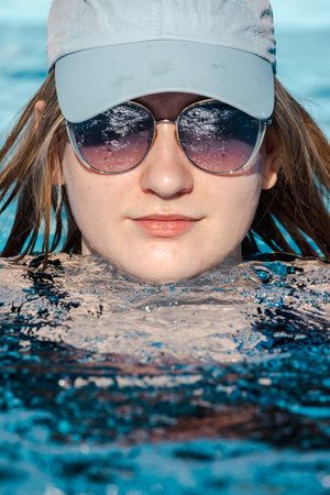 Teenage girl floating in a refreshing pool, wearing sunglasses and a light blue cap, with a calm expression and water reflections in her lensesの写真素材