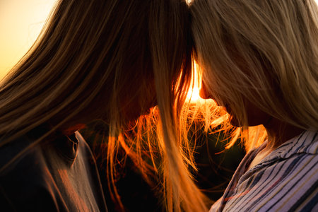 Tender sunset moment between mother and teenage daughter touching foreheads, their faces glowing in warm evening light and surrounded by soft strands of hairの写真素材