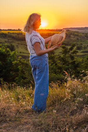 Middle-aged woman in striped shirt and jeans standing in a grassy field at sunset, holding a straw hat and enjoying the peaceful countryside viewの写真素材