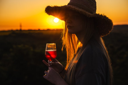 Teenage girl wearing a straw hat holds a glass of red drink and looks at the sunset in a peaceful countryside settingの写真素材