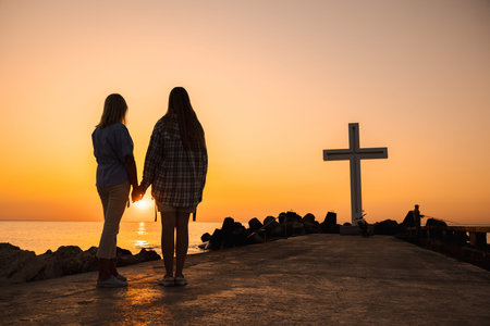 Mother and teenage daughter holding hands at sunrise near a large cross by the sea, silhouette view