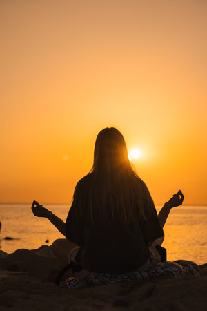 Young woman meditating in lotus position on seaside rocks at sunrise, hands in mudra, embracing...