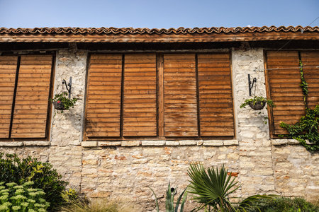 Old stone building with wooden shutters, tiled roof, and hanging flower baskets, surrounded by greenery and plants.の写真素材