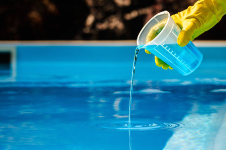 Yellow gloved hand pouring blue pool chemical from transparent measuring cup into water for...