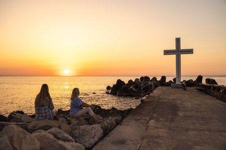 Two women sitting on rocks near a large cross by the sea at sunset in Varna, Bulgaria
