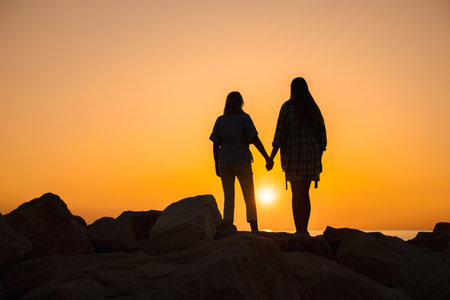 Silhouette of a mother and daughter holding hands on rocky shore at sunrise, symbolizing family love, emotional well-being, inner harmony, and a healthy lifestyle by the seaの写真素材