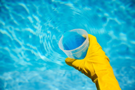 Yellow gloved hand holding transparent measuring cup with blue chemical liquid over pool water...