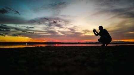 Silhouette of a man operating a drone controller by the lake at sunset with colorful evening skyの写真素材