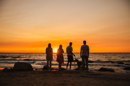Group of four people with a German Boxer dog standing on the sandy beach at sunset, gazing at the ocean horizon during a peaceful evening walk.の写真素材