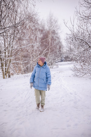 Elderly woman in a blue winter coat walking alone through a snowy countryside path. She looks up at the snow-covered trees around her, enjoying the calm and peaceful winter atmosphere.の写真素材