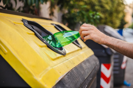 Close-up of a persons hand recycling a green plastic bottle into a yellow-lidded container for plastics on a street in Rome, promoting waste separation and environmental responsibility.の写真素材
