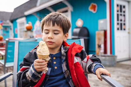 Little boy in a red winter jacket happily eating ice cream outside a colorful cafeの写真素材