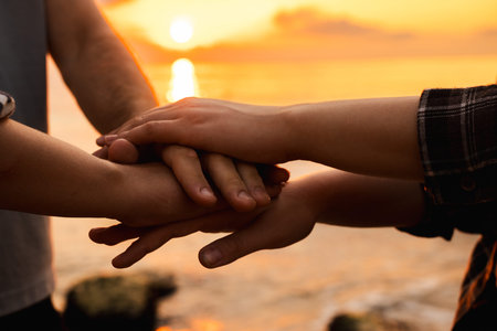 Close-up of three people joining hands in unity at sunrise on the beach, symbolizing teamwork,...