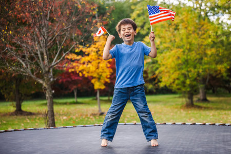 Smiling boy jumping high on trampoline outdoors holding Canada and USA flags in his hands on a...