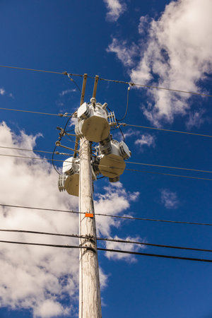 Wooden power pole with multiple electrical transformers and cables under a bright blue sky in the...