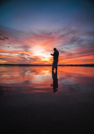 Man standing barefoot in shallow water during sunset, as the colorful sky reflects vividly on the calm lake surface.の写真素材