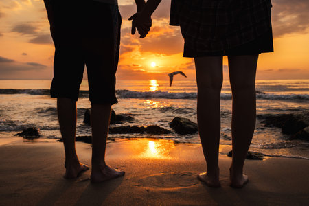 Couple holding hands on sandy beach at sunrise, watching a seagull fly over the glowing sea wavesの写真素材